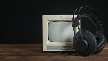 Black headphones with wire and retro monitor on wooden table on black background.