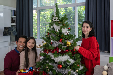 Asian happy family, father, mother, daughter help to prepare and gift or decorate Christmas tree in living room that decorated for christmas festival holiday concept