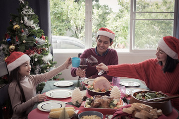 Asian family father, mother and daughter hold glass, they celebrate and cheers on Christmas day, they are happy with smile and have meal together in dining room that decorated with Christmas theme