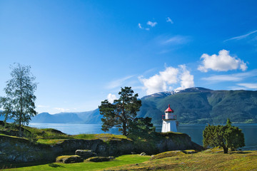 Fototapeta na wymiar White lighthouse tower with red top and nearby evergreen tree along the mountain lined shore of a Norwegian Fjord on a sunny day, with blue sky and fluffy white clouds.