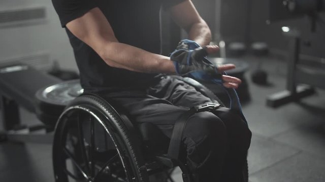 Workout, young disabled athlete in wheelchair puts on training gloves on his hands, training day in gym, people with disabilities.