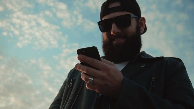 Stylish brutal man in black glasses and with a beard looks at the phone and laughs on a background of blue sky, slow motion, fashionable