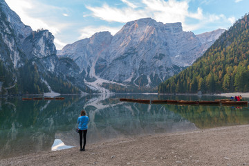 Fototapeta na wymiar A woman standing beside the lake Braies (Lago di Braies) in a morning with reflection of the mountain peak and boats on the calm lake in Dolomites, Italy