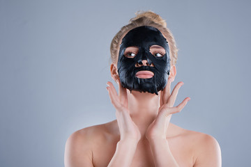 girl with naked shoulders posing at grey background with facial mask black, skin care concept, beauty photo, skin treatment, hydrating skin mask, smiling.