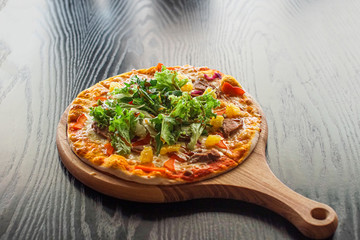 Fresh pizza with meat, pineapples and salad served on a board on black wooden table