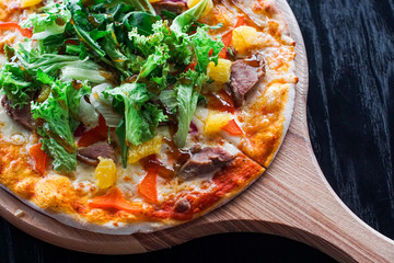 Top view of fresh pizza with meat, pineapples and salad served on a board on black wooden table