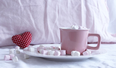 Fototapeta na wymiar Valentines composition. Cup of hot chocolate with Marshmallows, on the bed. Small red heart on background.Breakfast concept.