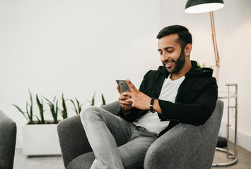 Trendy handsome Gen z Indian arabic man typing on mobile phone, sitting in waiting room before interview