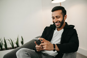 Trendy handsome Gen z Indian arabic man typing on mobile phone, smiling and laughing