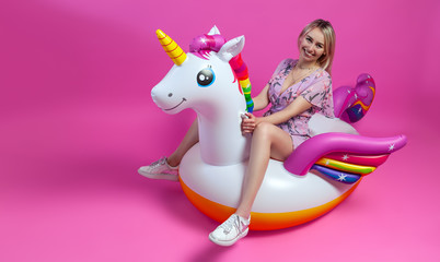 A beautiful blonde girl in a sexy sundress with slim legs in white sneakers sits on an inflatable multi-colored unicorn on a pink isolated background. Summer vacation at the beach and travel.