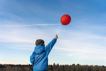 A boy in a winter blue jacket and hat stands with a red balloon on a stick against a blue clear sky with clouds. In the sky a trace from a flying airplane. The concept of innocence, dreams and hopes