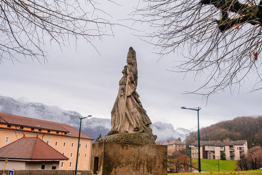 Sculpture of a woman against the backdrop of the high Alps in a gray cloudy sky f Annecy France. Photography of a stone sculpture near town houses, green grass, forest and mountains, autumn view.