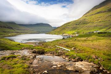 Fototapeta na wymiar Lake mirror near small town and green mountains covered with fluffy clouds, Faroe Islands.