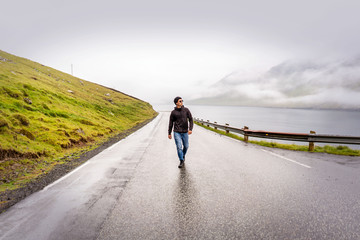 Obraz premium Young traveller walking y empty road after rain, green mountains, fjord and overcast sky on horizon. Faroe Islands, Denmark.