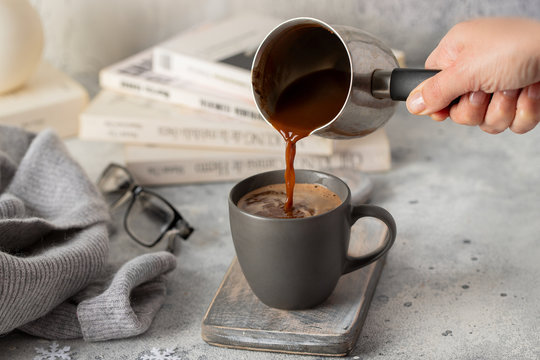 Hand Pouring Hot Chocolate Into The Cup For Relax Time At Home. Mug With Hot Drink And Books For Hygge Style.