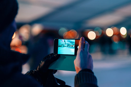 male hands holding a mobile phone on photo camera mode - man taking picture of a ice skating rink by night