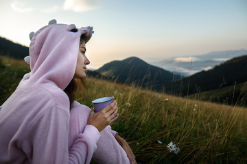 Young woman in a pink dragon costume drinks a hot drink from a cup and enjoys the scenery in the mountains