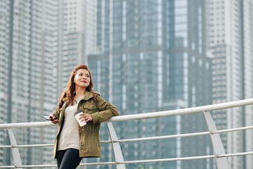 Positive pretty Asian woman with smartphone in hand leaning on bridge railing drinking coffee and enjoying beautiful view