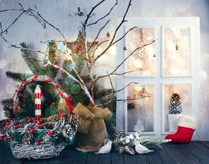 Obraz premium Christmas eve background with window, wicker festive basket, birds, candle, fir tree, pine, spruce cones, acorns on shabby wooden old table, New Year night winter holiday home natural material decor