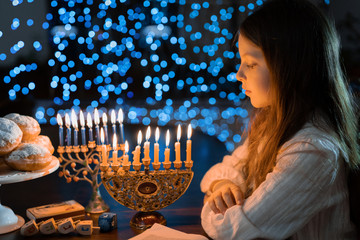 Child girl Looking at Menorah Candles on wooden table and sufganiyot on background light glitter bokeh overlay. Hanukkah jewish holiday Israel hebrew traditional family celebration invitation design