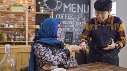 muslim woman customer give credit card to male waiter show bill and receipt in black leather tray in cafe restaurant. Service and payment in coffee shop concept. islam female client pay in debit card - obrazy, fototapety, plakaty
