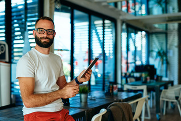 Fototapeta na wymiar Attractive bearded man with glasses sitting in a cafe searching on tablet 