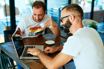 Attractive bearded man with glasses in a cafe working on laptop and chatting with a friend	