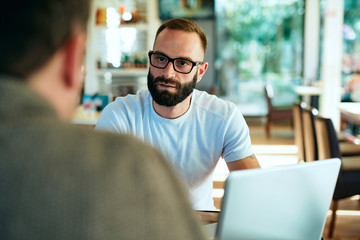 Fototapeta premium Attractive bearded man with glasses in a cafe working on laptop and chatting with a friend 