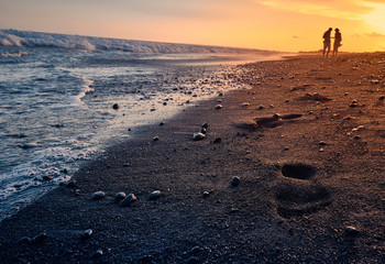 Footprints on sand with sunrise and silhouette of couple on background. Keramas black sand beach at sunset. Bali, Indonesia.