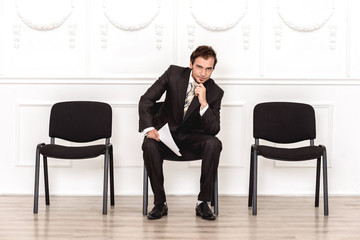 Job interview. Man sitting isolated on white waiting with resume touching chin smiling playful
