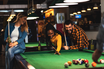 Four smiling friends in bar playing billiard together. - obrazy, fototapety, plakaty
