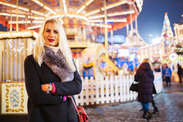 Image of young woman in coat with fur collar in park on background of carousel