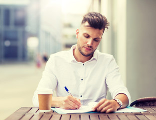 business, education and people concept - young man with coffee and folder writing at city street cafe