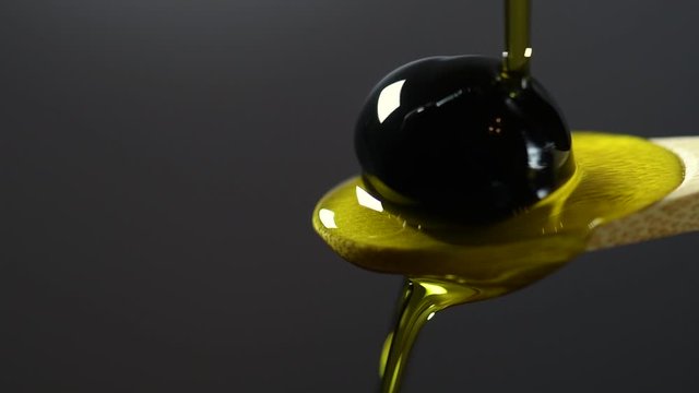 Olive oil is poured onto an olive lying on a spoon. Slow motion. Close-up of black olives on a wooden spoon on a gray background