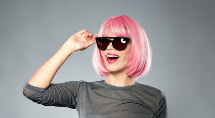 style, fashion and people concept - happy young woman in pink wig and black sunglasses over grey background