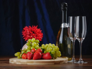 Fresh green grapes and red strawberry on a wooden board, Two champagne flutes  and a bottle of champagne without labell, flower in the background, Dark background. Selective focus.