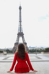 Young woman with long brown hair in red dress sitting back and look at Eiffel tower