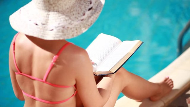 woman with hat reading a book by the pool.Relaxing in an outdoor swimming pool in the sun