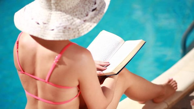 woman with hat reading a book by the pool.Relaxing in an outdoor swimming pool in the sun