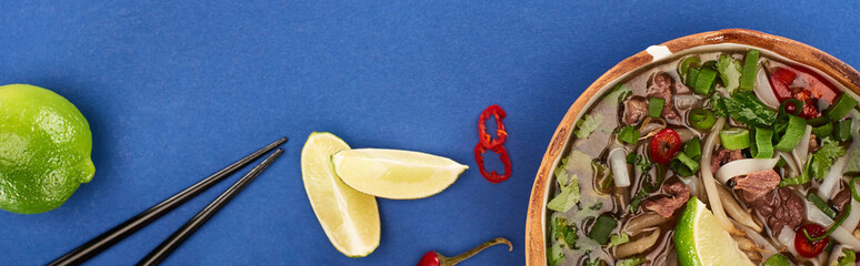 top view of pho in bowl near chopsticks, lime, chili on blue background, panoramic shot