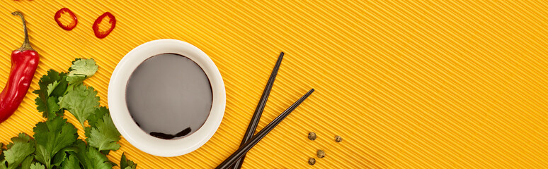 top view of chili, soy sauce and coriander near chopsticks on yellow background, panoramic shot