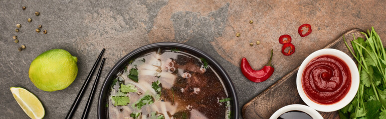 top view of pho in bowl near chopsticks, lime, chili and soy sauces and coriander on stone...