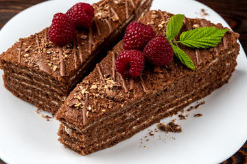 Slice of chocolate cake with milk filling and raspberry on white plate on dark wooden background.