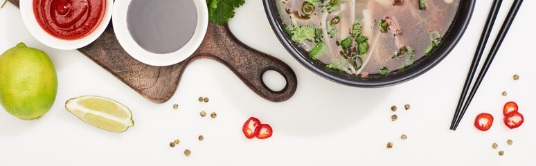 top view of pho in bowl near chopsticks, lime, chili and soy sauces and coriander on white background, panoramic shot