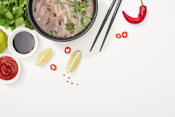 top view of pho in bowl near chopsticks, lime, chili and soy sauces and coriander on white...