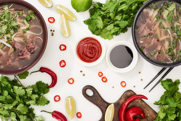 top view of pho in bowls near chopsticks, lime, chili and soy sauces and coriander on white...
