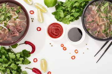 top view of pho in bowls near chopsticks, lime, chili and soy sauces and coriander on white background