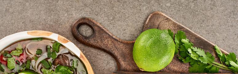 top view of pho in bowl near lime and coriander on wooden cutting board on grey background,...