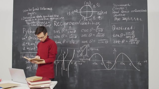 Lockdown of young Asian man wearing casual clothes walking with book in his hands, looking in it and then looking at blackboard