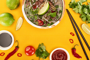 top view of pho in bowl near chopsticks, lime, chili and soy sauces and coriander on yellow...